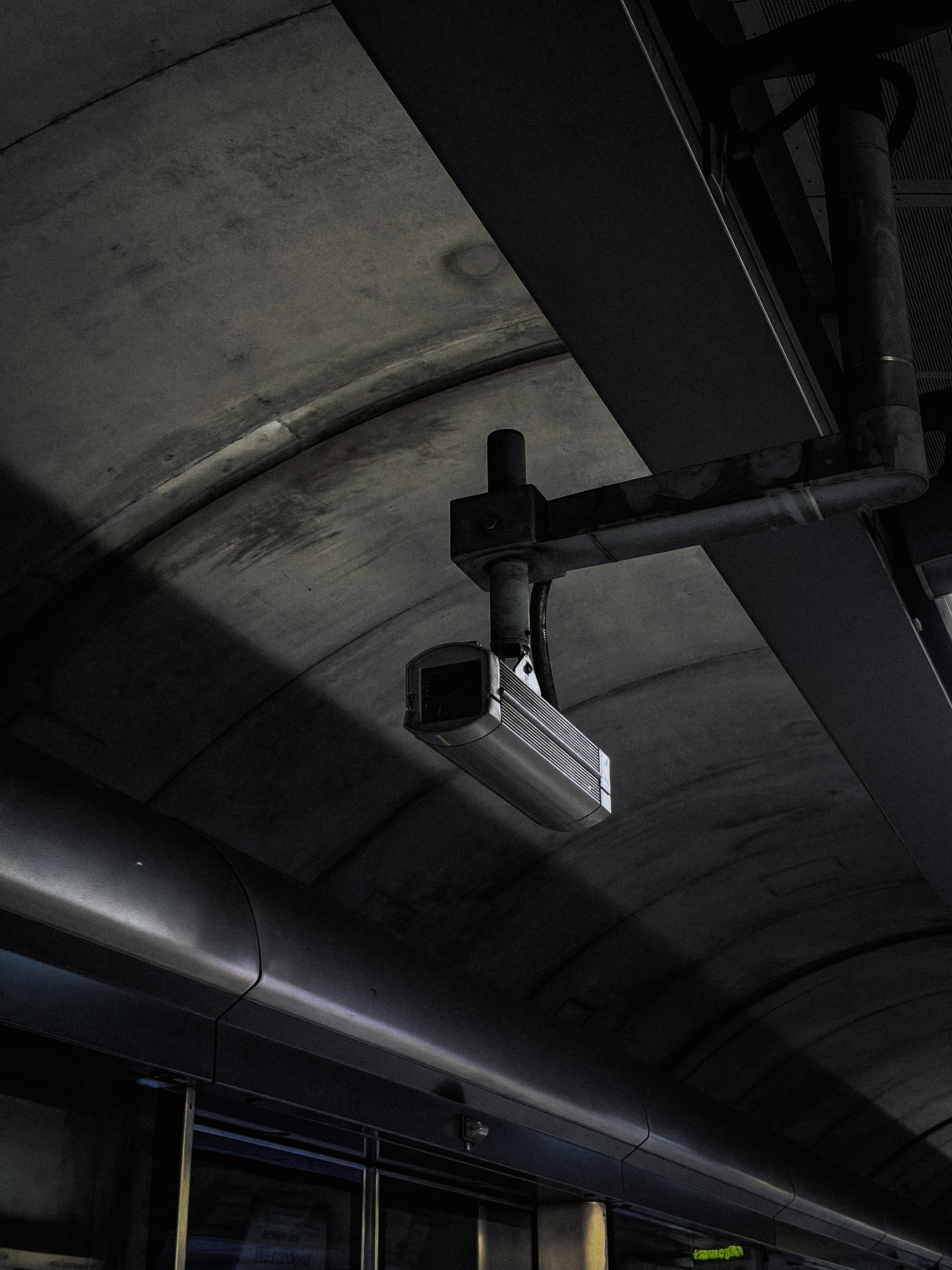 A CCTV camera suspended beneath a curved concrete ceiling, framed by dark steel beams and pipework