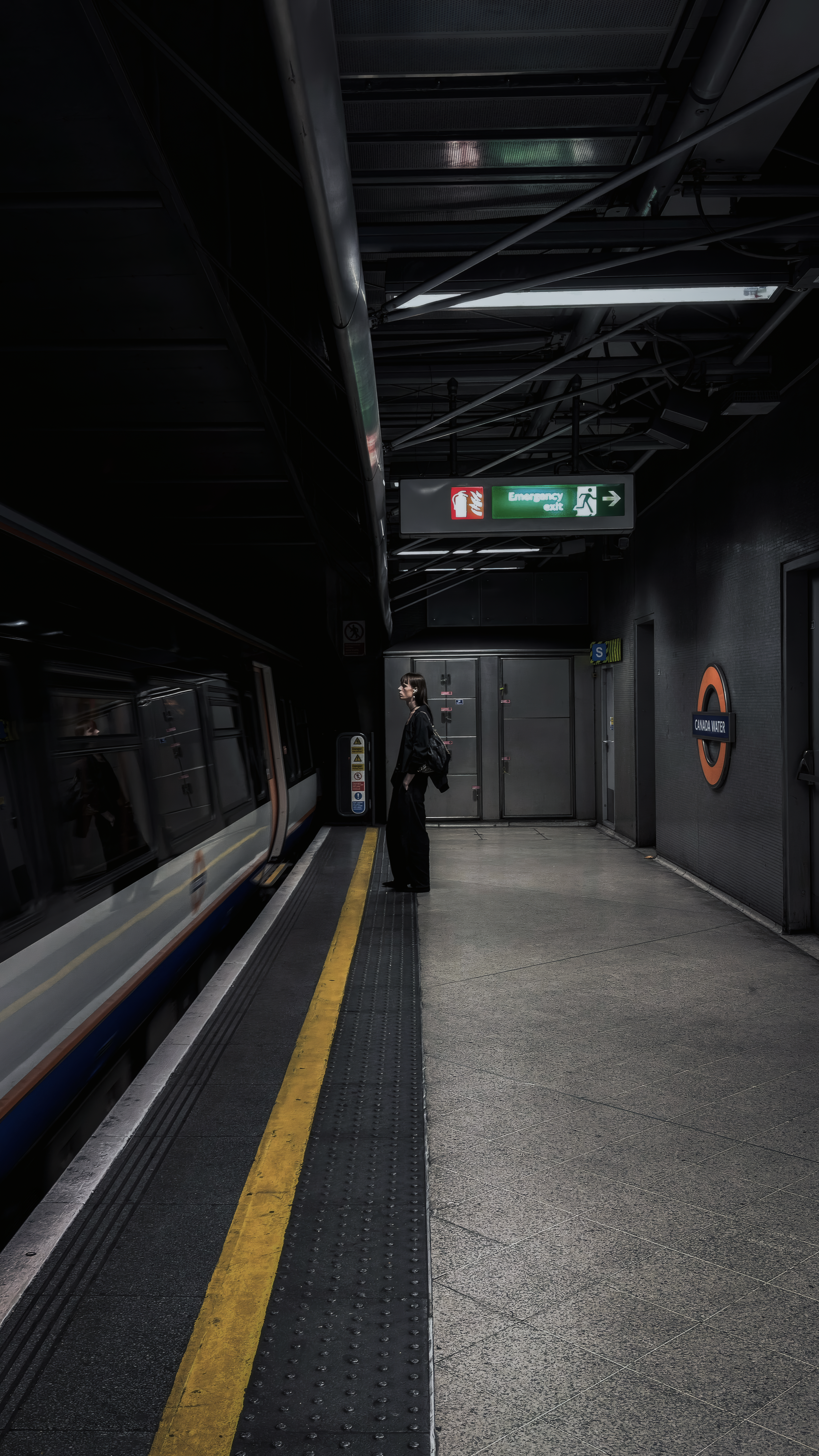 A lone commuter beside a passing train and yellow platform line at Canada Water