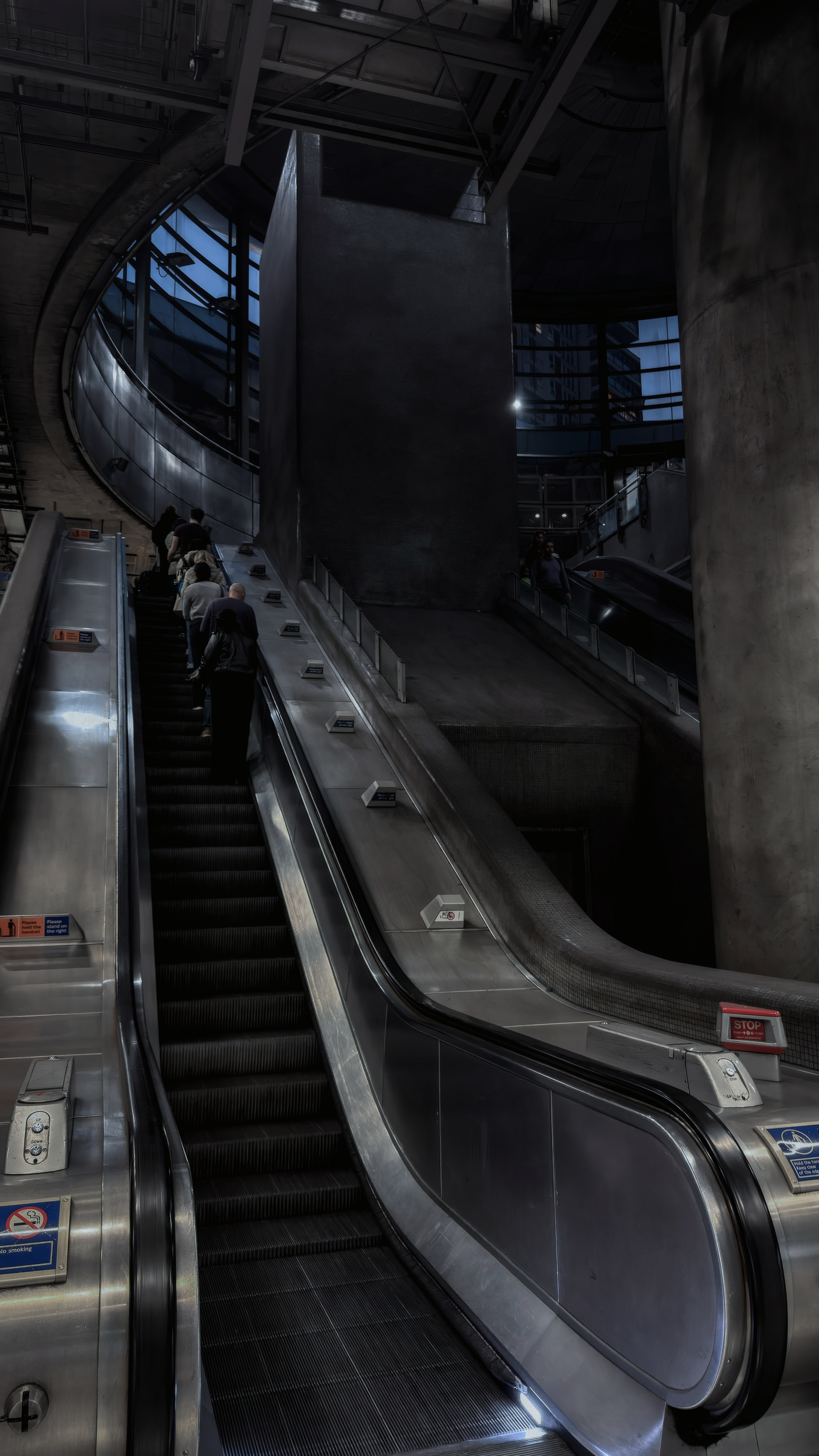 Sweeping steel escalator rails beneath concrete columns and blue light at Canada Water station