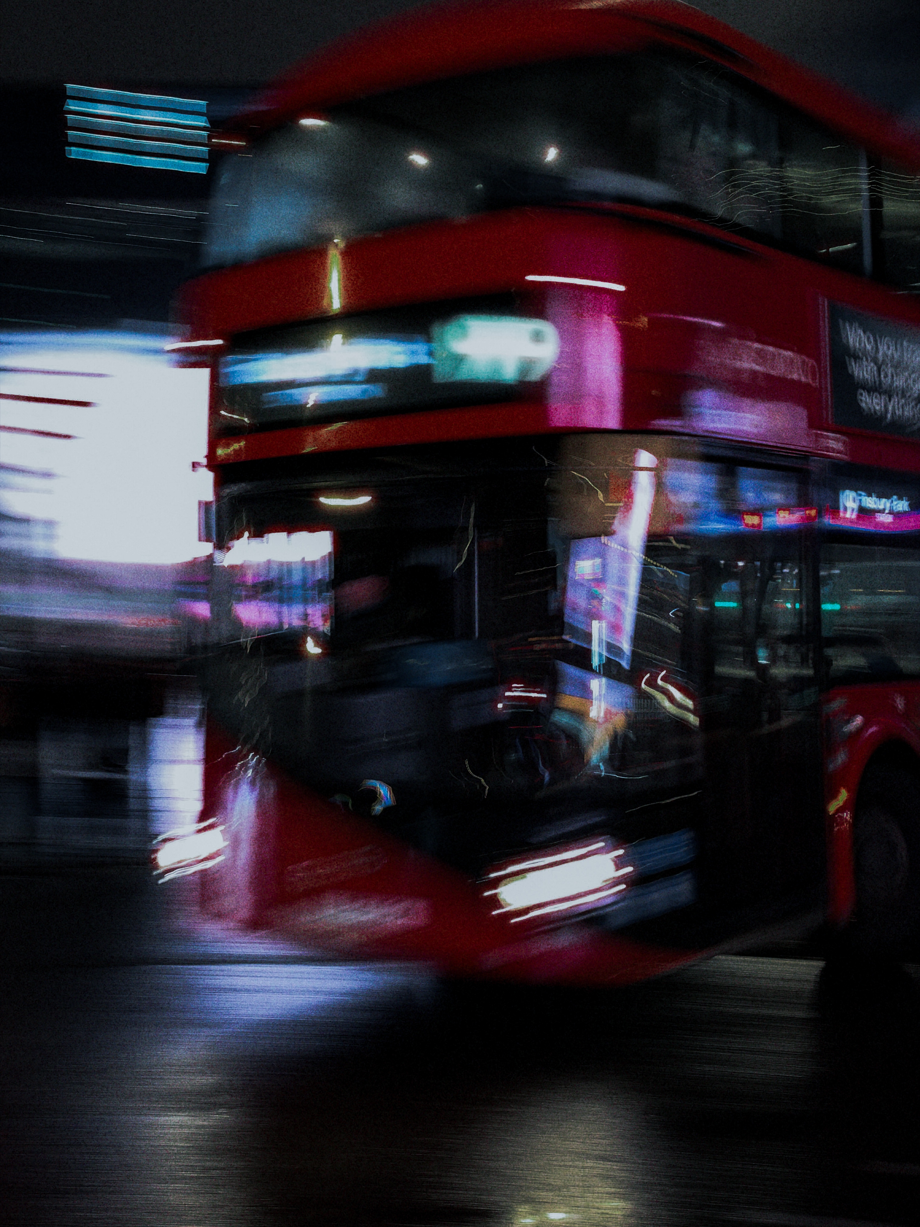 A red double-decker bus turning through a dark street with blurred lights and wet-road reflections